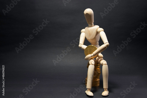 Model of a wooden figure sitting on a pile of coins being happy, holding a coin in his arms on a black background. Business concept