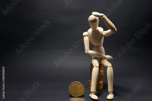 Model of a wooden figure sitting on a pile of coins being confused, holding a coin in his arms on a black background. Business concept