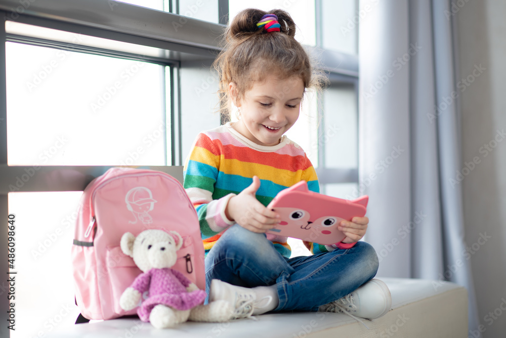 Positive little girl, holding tablet computer in her hands. Advertising new gadget, educational app. The girl sits near the window. She is wearing a colored T-shirt. Games.Children use technology. Positive little girl, holding tablet computer in her hands. Advertising new gadget, educational app. The girl sits near the window. She is wearing a colored T-shirt. Games.Children use technology.