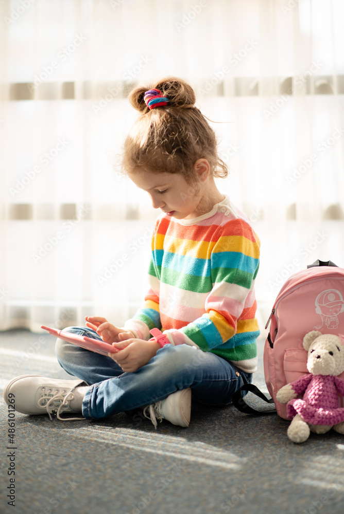 Positive little girl, holding tablet computer in her hands. Advertising new gadget, educational app. The girl sits near the window. She is wearing a colored T-shirt. Games.Children use technology. Positive little girl, holding tablet computer in her hands. Advertising new gadget, educational app. The girl sits near the window. She is wearing a colored T-shirt. Games.Children use technology.