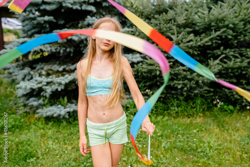 Happy preteen girl playing with gymnastic ribbon outdoor. Young lady with gymnastic tape played in a summer park or backyard. She is dressed in a green short top and shorts, colorful ribbon.