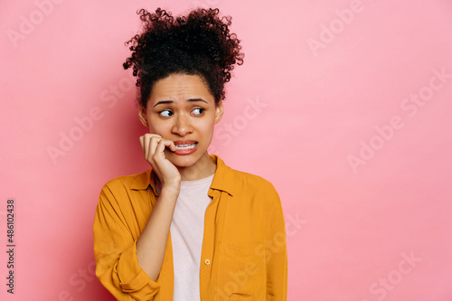 Stunned african american young woman, in casual wear, looking stressed and nervous with hands on mouth biting nails, looking aside, is going through, stands on isolated pink background