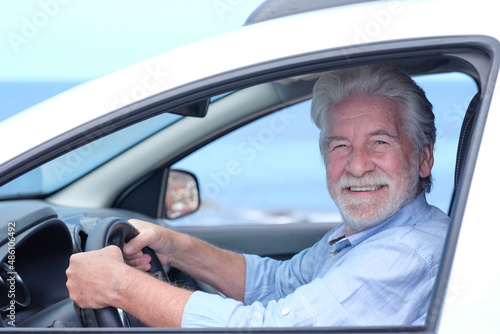 Beautiful adult active white-haired senior man sitting inside the car touching the steering wheel looking at camera smiling. Elderly grandparent with white beard enjoying travel. Horizon over water © luciano