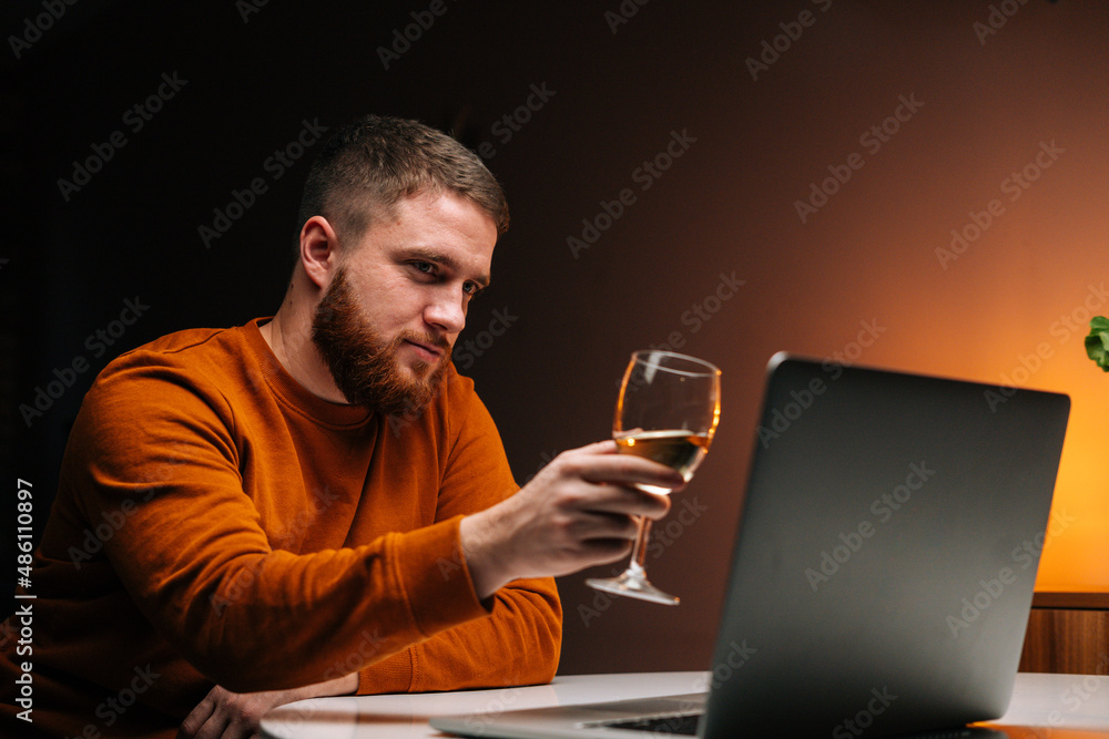 Close-up of young man enjoying distant dating using laptop, clinking glass with champagne, talking celebration toasting via video call, sitting at table with candle in dark room, looking on screen. Close-up of young man enjoying distant dating using laptop, clinking glass with champagne, talking celebration toasting via video call, sitting at table with candle in dark room, looking on screen.