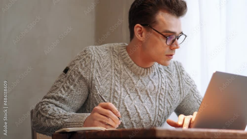 Smiling man wearing eyeglasses writing something from laptop to notepad at home Smiling man wearing eyeglasses writing something from laptop to notepad at home