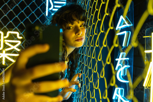 A young woman with curly hair takes a selfie near a yellow metal grid and in neon lighting. photo