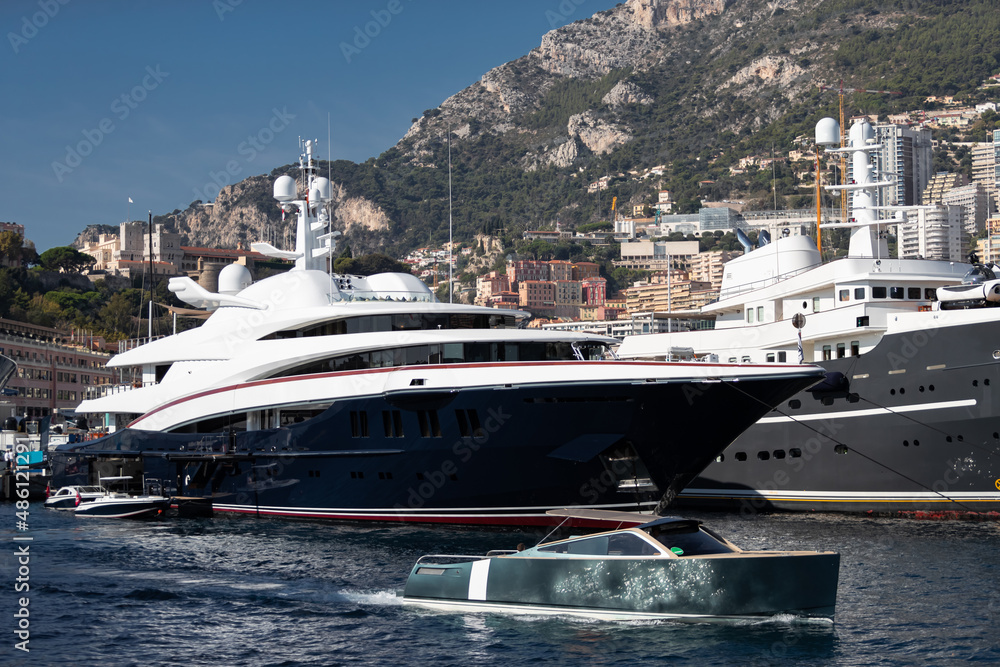 A lot of huge yachts are in port of Monaco at sunny day, Monte Carlo, mountain is on background, glossy board of the motor boat, megayachts are moored in marina, sun reflection on glossy board