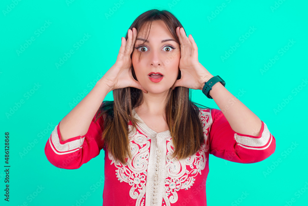 Fototapeta premium young beautiful moroccan woman wearing traditional caftan dress over green background with scared expression, keeps hands on head, jaw dropped, has terrific expression. Omg concept