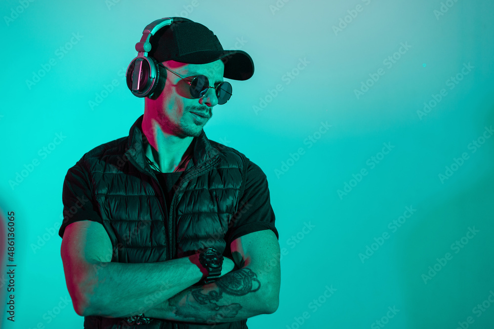 Hipster man model with sunglasses, cap and headphones in fashion black clothes in studio with cyan and pink color. Guy listening to music