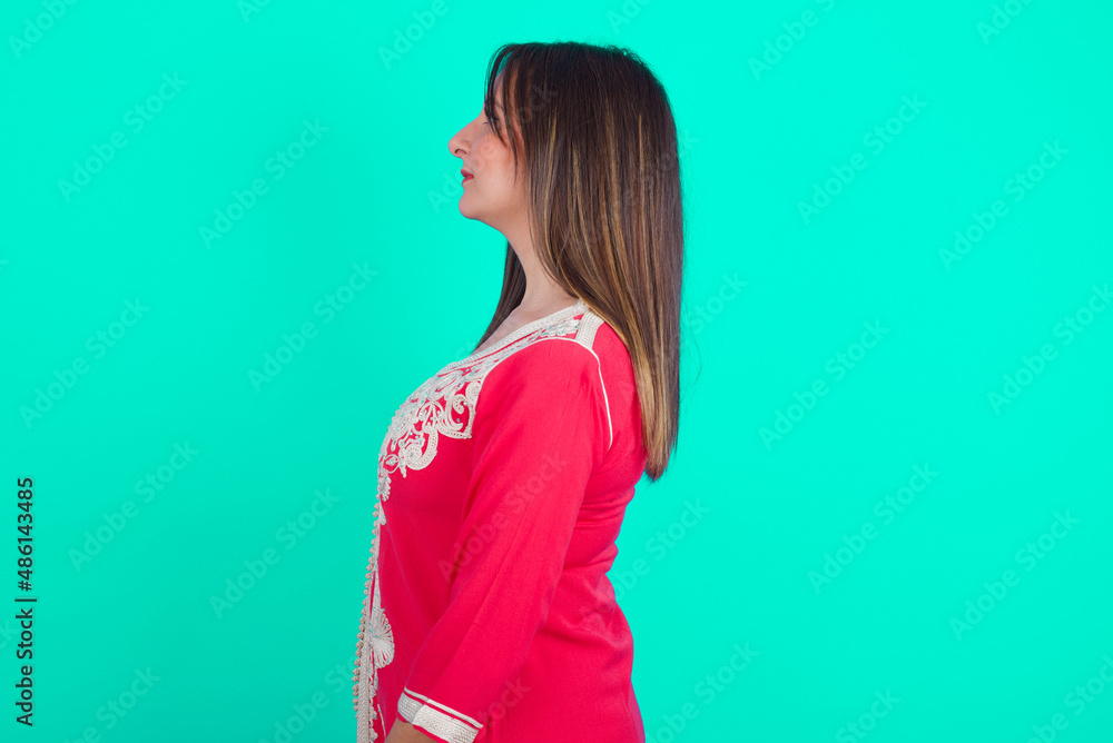 young beautiful moroccan woman wearing traditional caftan dress over green background looking to side, relax profile pose with natural face with confident smile. young beautiful moroccan woman wearing traditional caftan dress over green background looking to side, relax profile pose with natural face with confident smile.