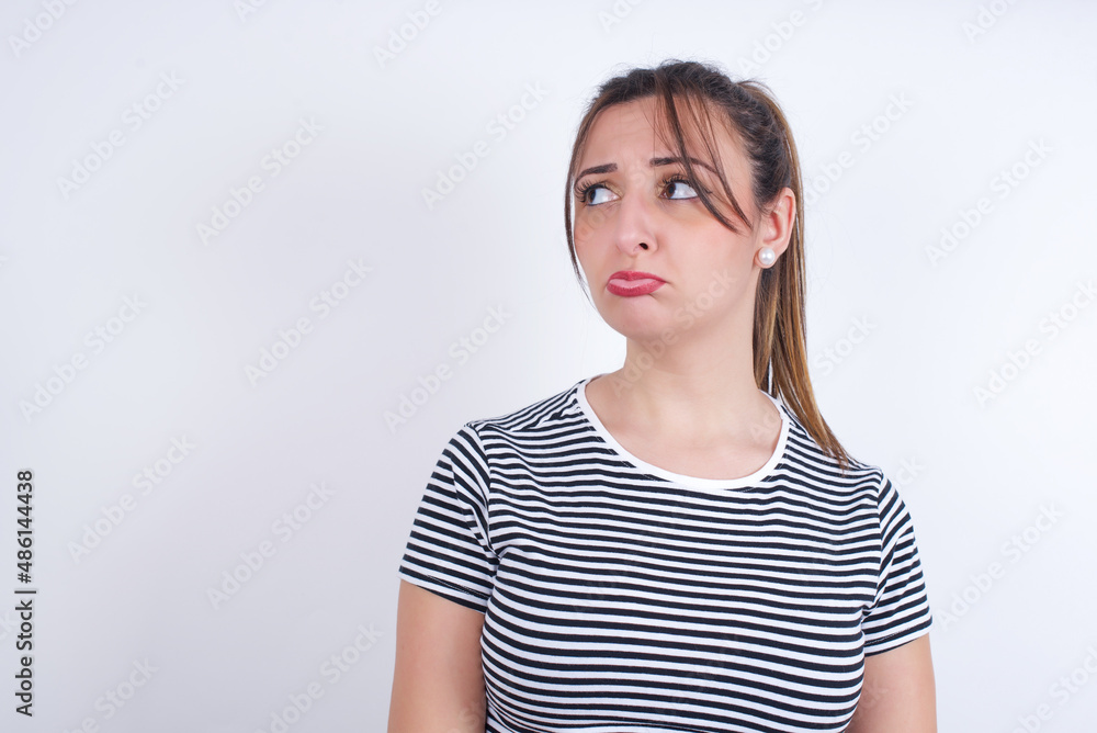 Dissatisfied young Arab woman wearing striped t-shirt over white background purses lips and has unhappy expression looks away stands offended. Depressed frustrated model. Dissatisfied young Arab woman wearing striped t-shirt over white background purses lips and has unhappy expression looks away stands offended. Depressed frustrated model.
