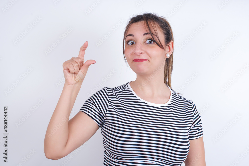 young Arab woman wearing striped t-shirt over white background purses lip and gestures with hand, shows something very little. young Arab woman wearing striped t-shirt over white background purses lip and gestures with hand, shows something very little.