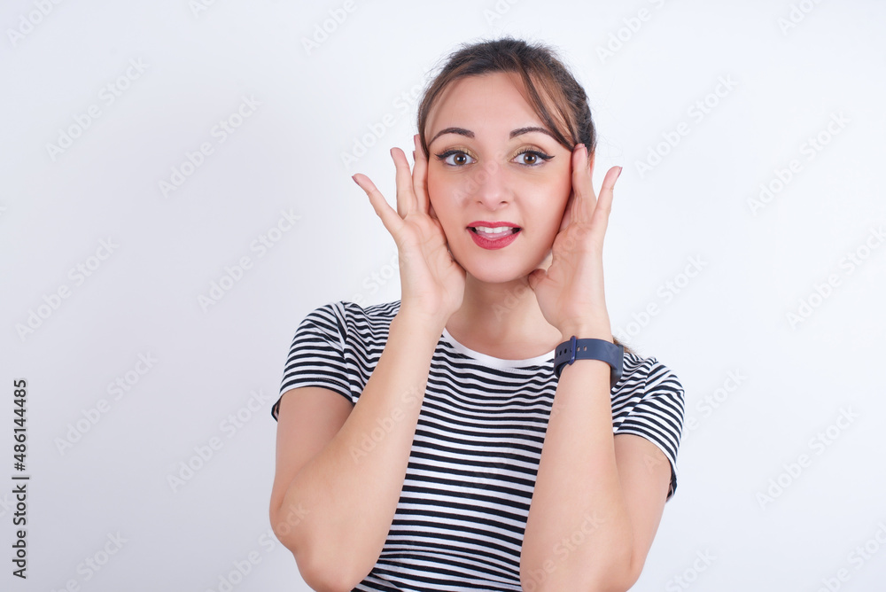 Happy young Arab woman wearing striped t-shirt over white background touches both cheeks gently, has tender smile, shows white teeth, gazes positively straightly at camera, Happy young Arab woman wearing striped t-shirt over white background touches both cheeks gently, has tender smile, shows white teeth, gazes positively straightly at camera,