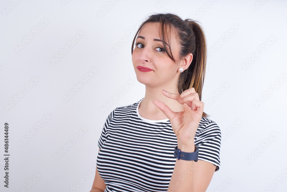 Displeased young Arab woman wearing striped t-shirt over white background shapes little hand sign demonstrates something not very big. Body language concept. Displeased young Arab woman wearing striped t-shirt over white background shapes little hand sign demonstrates something not very big. Body language concept.