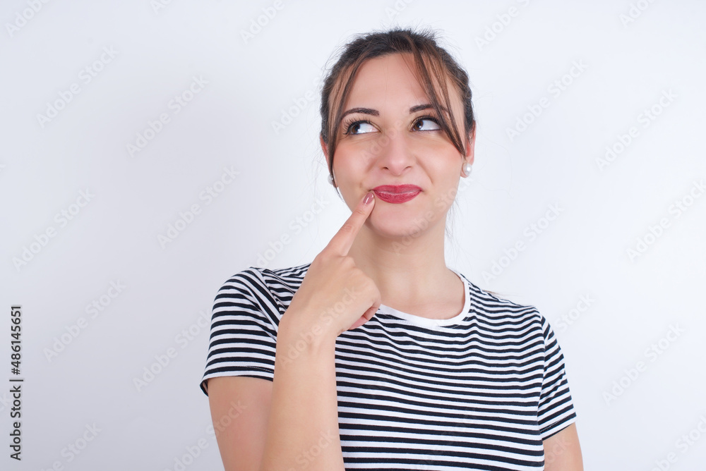 Lovely dreamy young Arab woman wearing striped t-shirt over white background keeps finger near lips looks aside copy space. Lovely dreamy young Arab woman wearing striped t-shirt over white background keeps finger near lips looks aside copy space.