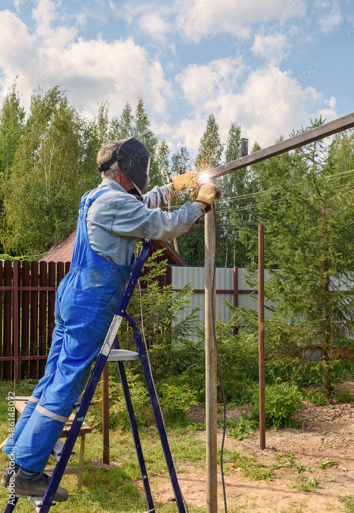 Man welder in a welding mask, construction uniform and protective gloves cooks metal on a street construction site. Construction of a pavilion, pergola near a country house on a summer day. Man welder in a welding mask, construction uniform and protective gloves cooks metal on a street construction site. Construction of a pavilion, pergola near a country house on a summer day.