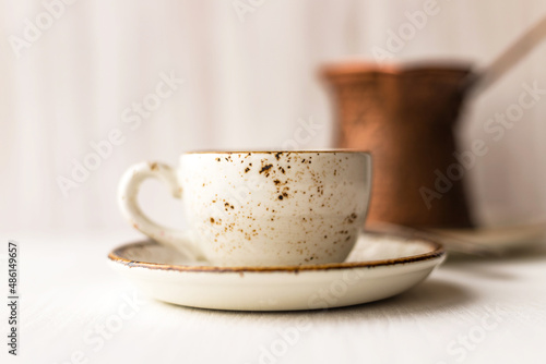 Cup of black Turkish coffee and coffee maker turk on white wooden table against wall background