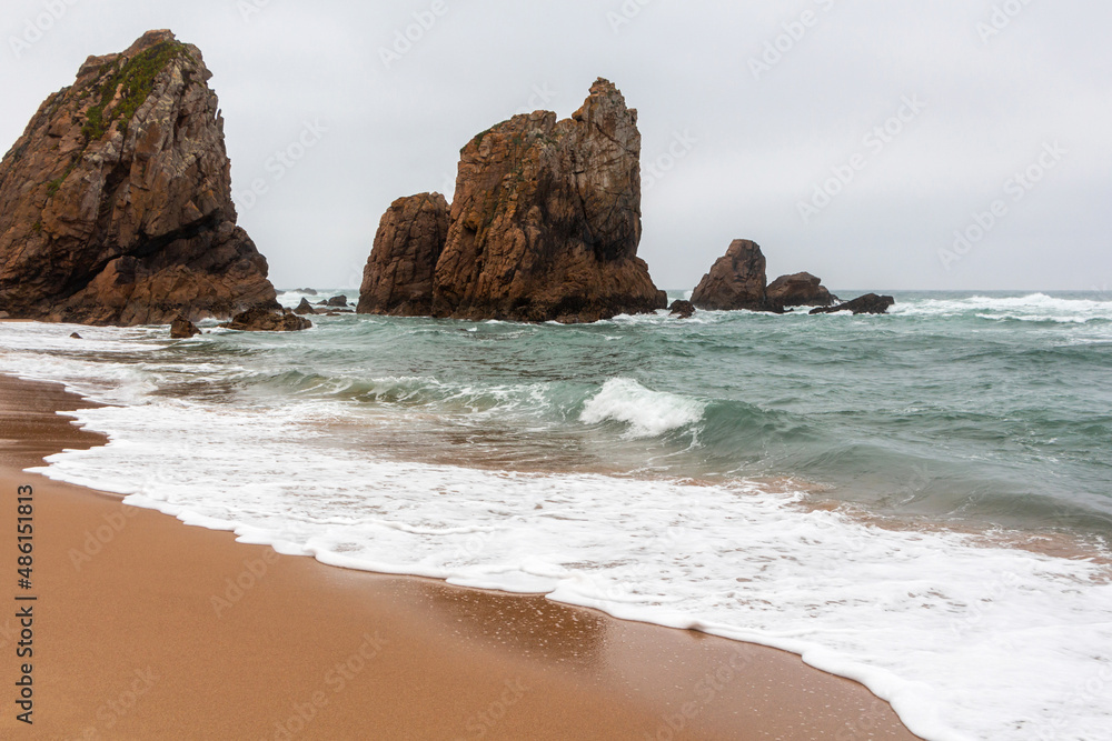 Beautiful landscape of the sandy beach of Ursa with an incoming foamy wave on the shore of the Atlantic Ocean with turquoise raging water among high rocks and stones, Portugal