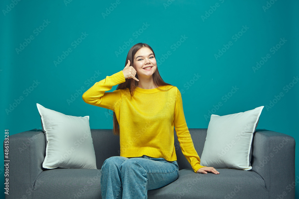 Beautiful smiling young girl showing call me gesture with fingers, suggest communication, sitting on couch Beautiful smiling young girl showing call me gesture with fingers, suggest communication, sitting on couch
