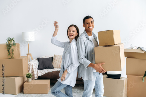 Cheerful happy marriage couple relocate their first own home. Spouses of different nationalities, standing in living room, guy holding a box, girl keys, look at camera, cardboard boxes on background