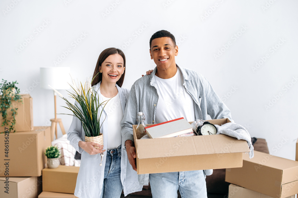Portrait of a happy couple in love, husband and wife of different nationalities, moved to their new home, the guy holds a box with things, the girl keys and a flowerpot, look at the camera, smile Portrait of a happy couple in love, husband and wife of different nationalities, moved to their new home, the guy holds a box with things, the girl keys and a flowerpot, look at the camera, smile