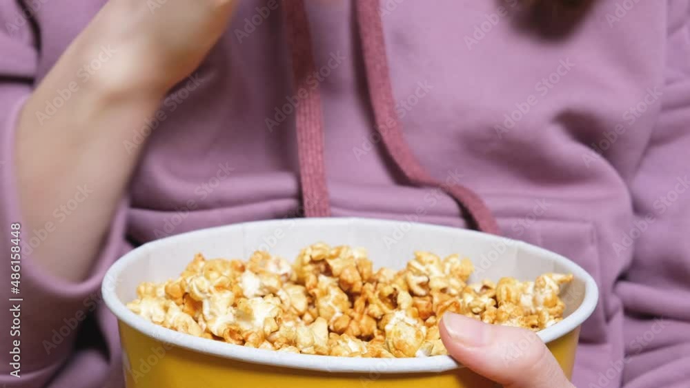 Close up view on female hand holding and eating a popcorn while watching a movie in the living room Close up view on female hand holding and eating a popcorn while watching a movie in the living room