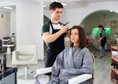 Positive male hairdresser making hair styling for young woman, working day in beauty salon