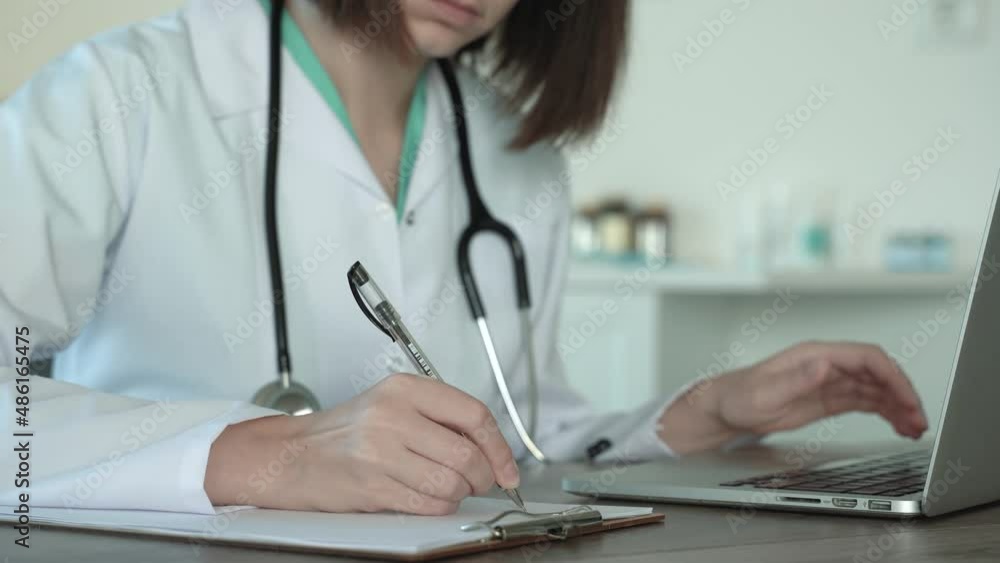 Close up view of female doctor and white coat using laptop in medical office, writes patient diagnosis prescription in medical notepad. Distance counsulting in covid 19 quarantine. 4k footage Close up view of female doctor and white coat using laptop in medical office, writes patient diagnosis prescription in medical notepad. Distance counsulting in covid 19 quarantine. 4k footage