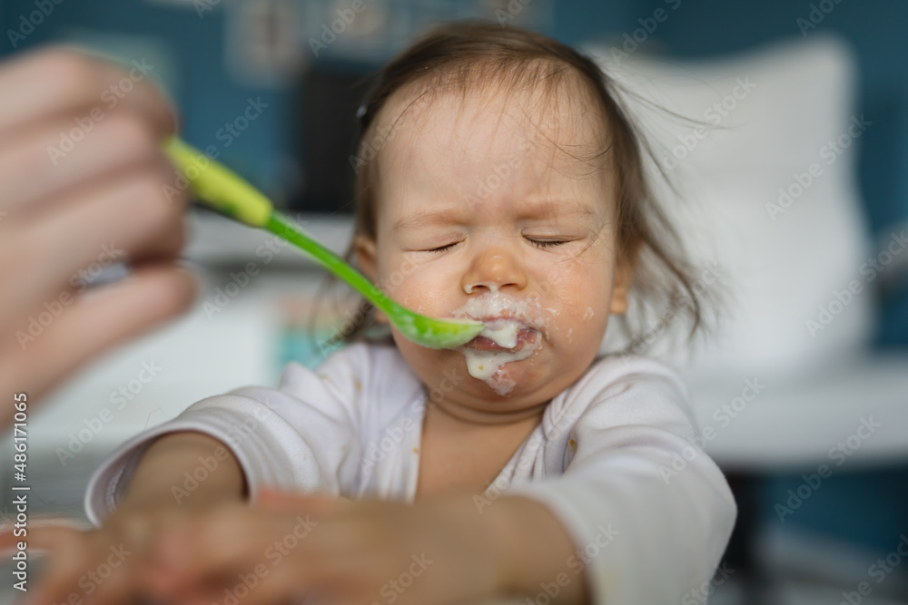 One baby close up on small caucasian girl and hand of her unknown mother feeding her at home in day small child with food around mouth making mess dirty while eating real people growing up copy space One baby close up on small caucasian girl and hand of her unknown mother feeding her at home in day small child with food around mouth making mess dirty while eating real people growing up copy space