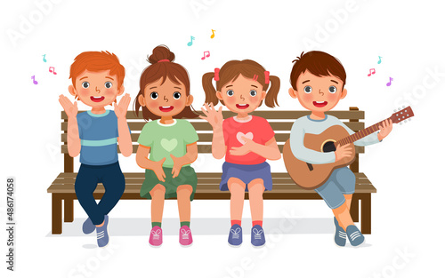 happy little boys and girls clapping hands, singing, playing guitar and having fun together sitting on bench