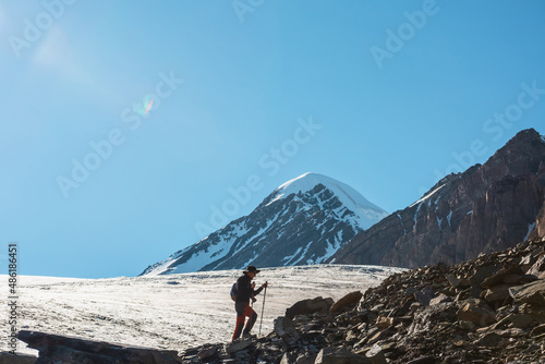 Scenic alpine landscape with silhouette of hiker with trekking poles against large glacier tongue and snow mountain peak in sunlight. Man with backpack in high mountains under blue sky in sunny day.