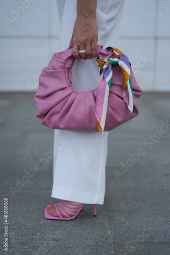 the leg of the blogger's fashionista girl in white trousers and pink sandals, the girl is holding a pink handbag with a scarf tied on it