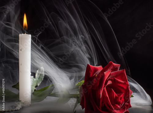 the bright flame of a burning candle illuminates a red rose lying nearby.