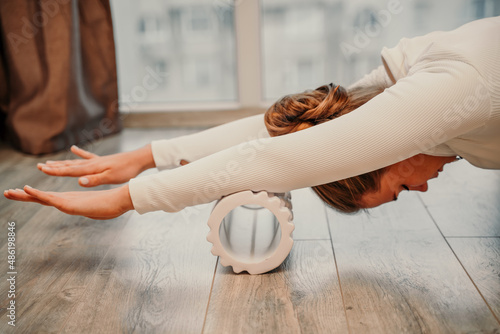Adult athletic woman, in white bodysuit, performing fascia exercises on the floor - caucasian woman using a massage foam roller - a tool to relieve tension in the back and relieve muscle pain - the photo
