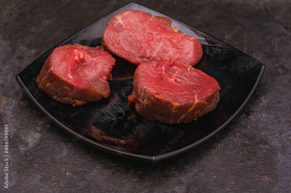 Three pieces of raw beef on a plate on dark background