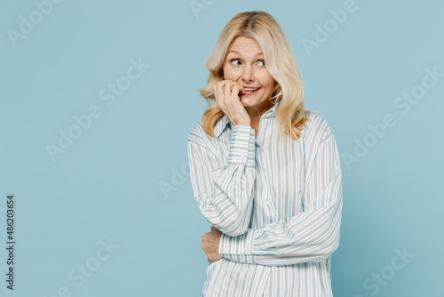 Elderly puzzled thoughtful puzzled confused woman 50s wear striped shirt biting nails fingers look aside isolated on plain pastel light blue color background studio portrait. People lifestyle concept.