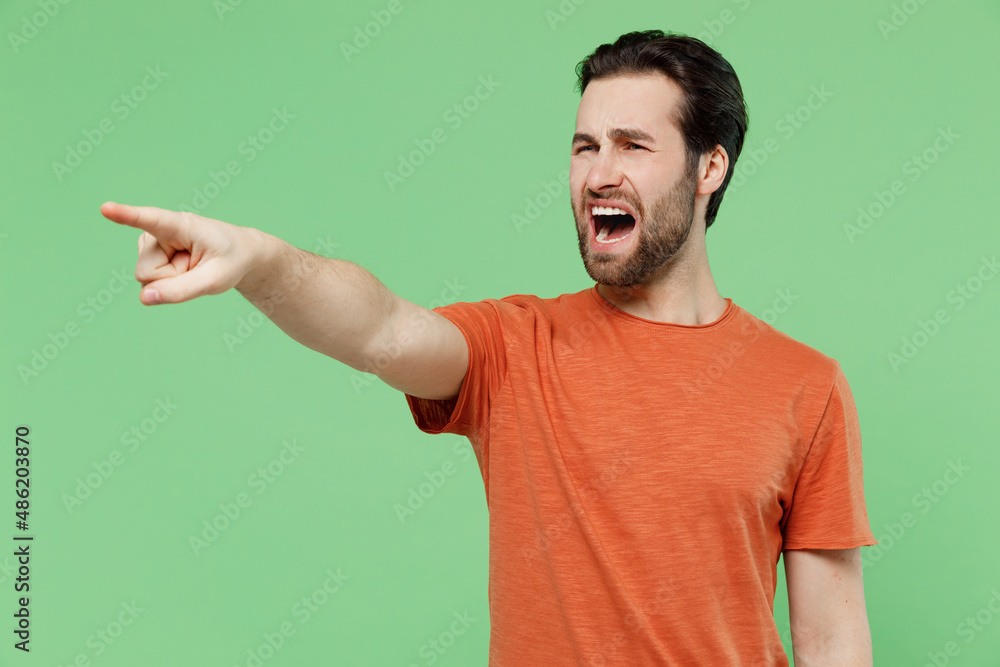 Young angry sad strict furious caucasian man 20s wear casual orange t-shirt point index finger aside scream isolated on plain pastel light green background studio portrait. People lifestyle concept. Young angry sad strict furious caucasian man 20s wear casual orange t-shirt point index finger aside scream isolated on plain pastel light green background studio portrait. People lifestyle concept.