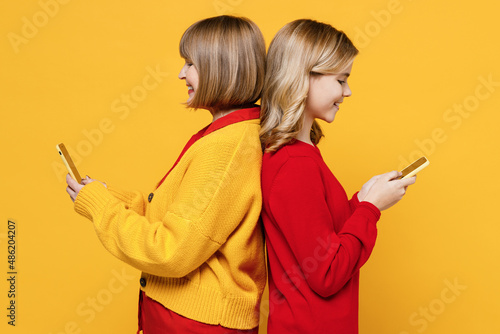 Side view woman 50s in red shirt have fun with teenager girl 12-13 years old. Grandmother granddaughter hold in hand use mobile cell phone isolated on plain yellow background Family lifestyle concept