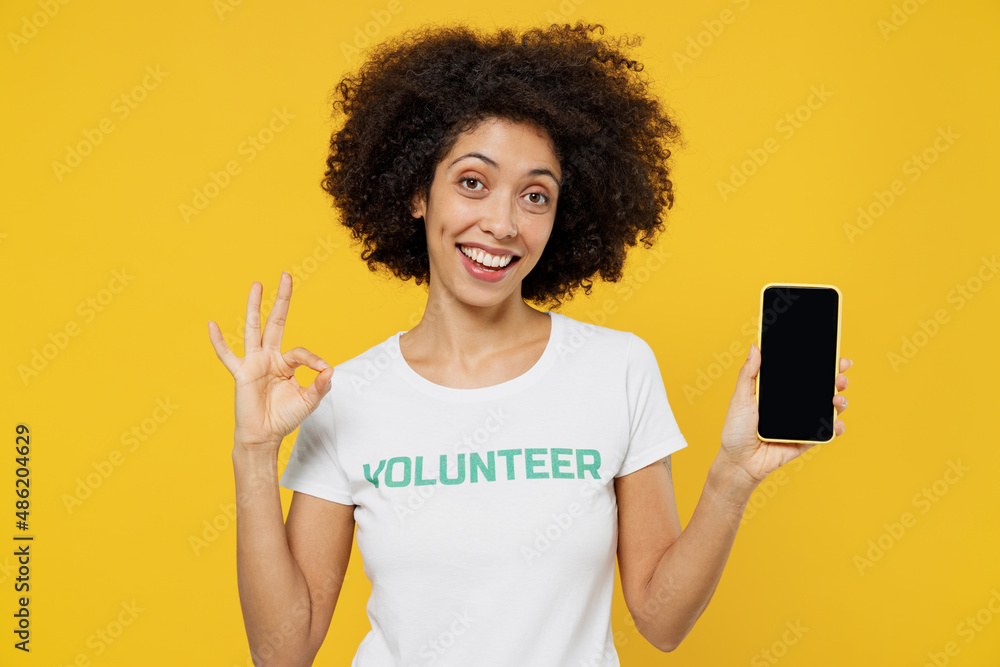 Young overjoyed fun woman of African American ethnicity wear white volunteer t-shirt hold use mobile cell phone with blank screen workspace area show okay gesture isolated on plain yellow background Young overjoyed fun woman of African American ethnicity wear white volunteer t-shirt hold use mobile cell phone with blank screen workspace area show okay gesture isolated on plain yellow background