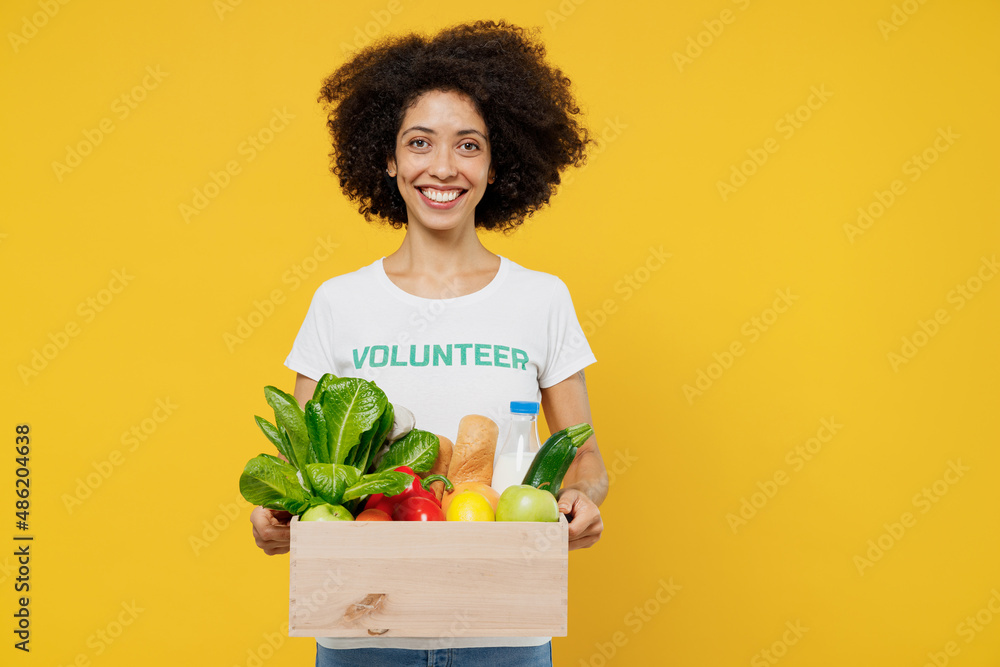 Young woman of African American ethnicity wears white volunteer t-shirt hold box with food vegetables isolated on plain yellow background. Voluntary free work assistance help charity grace concept Young woman of African American ethnicity wears white volunteer t-shirt hold box with food vegetables isolated on plain yellow background. Voluntary free work assistance help charity grace concept