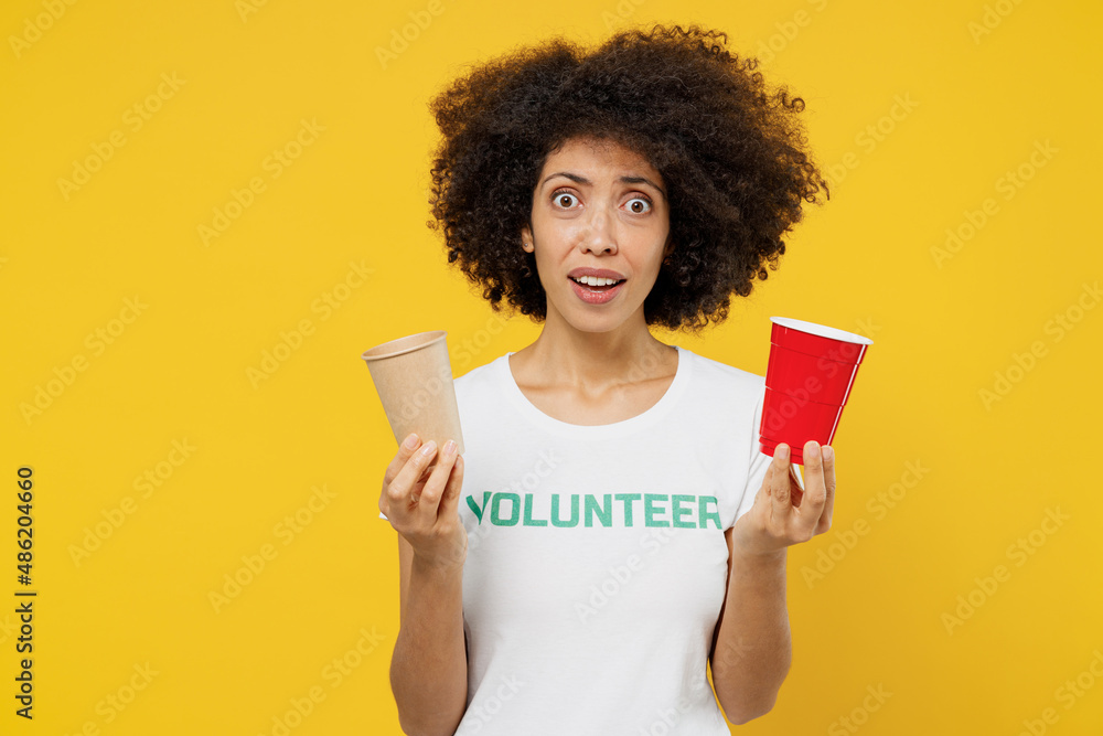 Young shocked woman of African American ethnicity in white volunteer tshirt hold choose paper plastic cup isolated on plain yellow background Voluntary free work assistance help charity grace concept Young shocked woman of African American ethnicity in white volunteer tshirt hold choose paper plastic cup isolated on plain yellow background Voluntary free work assistance help charity grace concept
