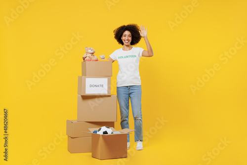 Full body young woman of African American ethnicity in white volunteer t-shirt hold boxes with presents waving hand isolated on plain yellow background. Voluntary free work assistance help concept photo
