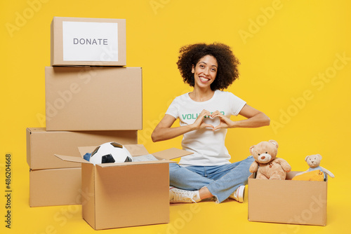 Full body young woman of African American ethnicity in white volunteer t-shirt sit near boxes with presents show heart gesture isolated on plain yellow background. Voluntary free work help concept. photo