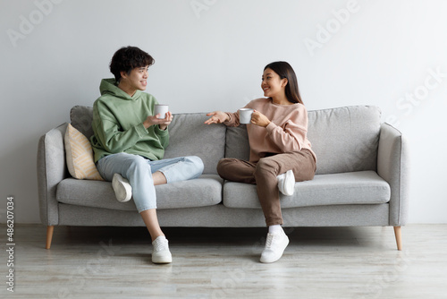 Young Asian man and woman drinking coffee and talking while sitting on couch at home