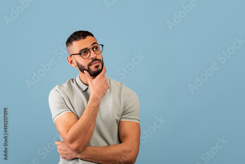 Pensive middle eastern guy thinking and touching chin, looking aside at free space, posing over blue background