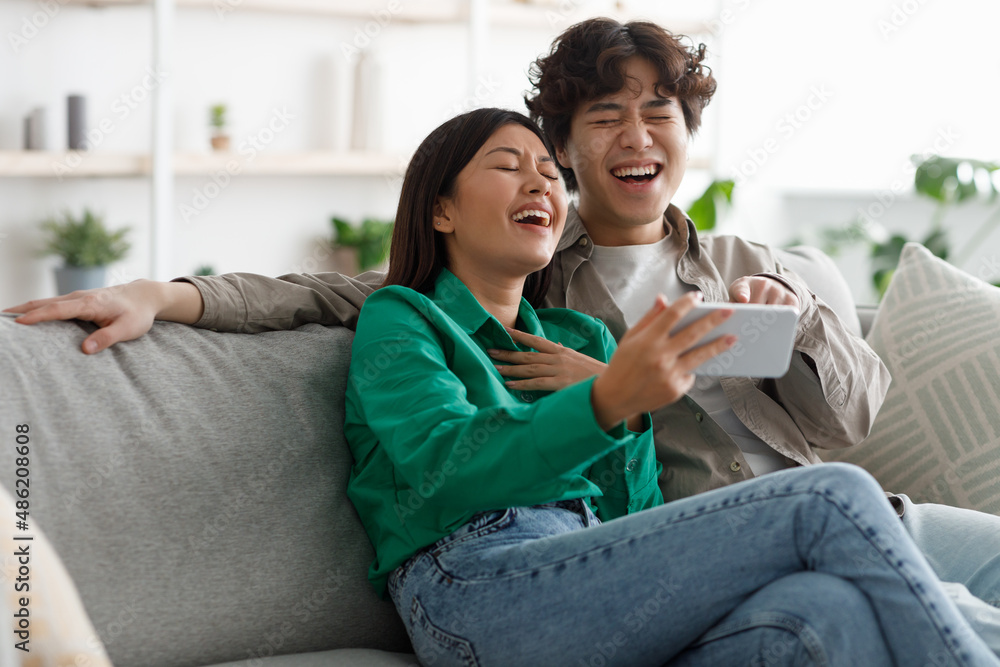 Joyful young Asian couple watching funny videos on smartphone, laughing together, relaxing on couch at home Joyful young Asian couple watching funny videos on smartphone, laughing together, relaxing on couch at home