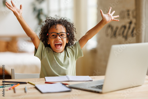 Little african american school boy raising hands up with excitement during home distance education photo