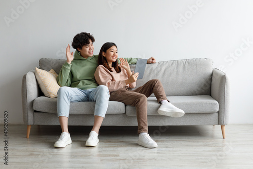 Cheerful millennial Asian couple using tablet for online communication, sitting on sofa, waving at webcam at home