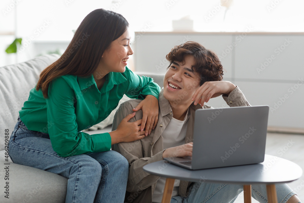 Happy millennial Asian couple using laptop, surfing web, watching movie, relaxing at home on weekend morning Happy millennial Asian couple using laptop, surfing web, watching movie, relaxing at home on weekend morning