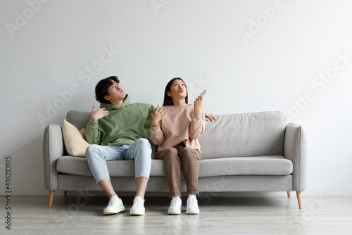 Young Asian couple suffering from summer heat, holding remote control, switching on air conditioner at home, mockup